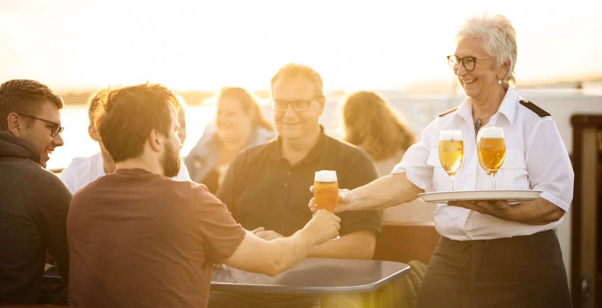 Eine Frau serviert Bier an eine Gruppe von Menschen auf einem Boot bei Sonnenuntergang.