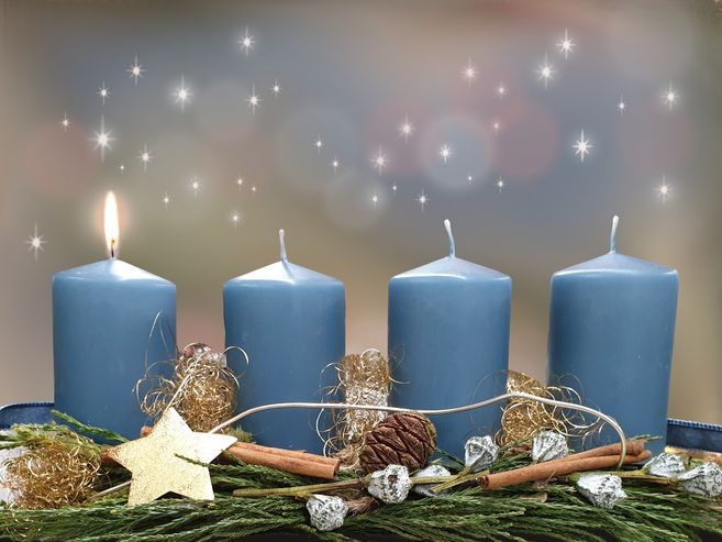 Ein Adventskranz mit vier blauen Kerzen, von denen eine brennt, umgeben von Tannenzweigen, Zimtstangen und goldenen Dekorationen.