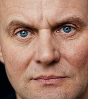 Nahaufnahme eines Mannes mit blauen Augen und ernstem Gesichtsausdruck vor schwarzem Hintergrund.