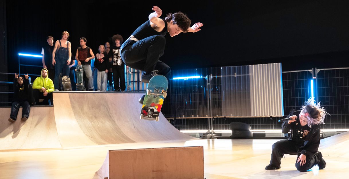 A skateboarder dynamically jumps over an obstacle while a performer with a microphone sings in a squatting position and a group of spectators watch in the background.