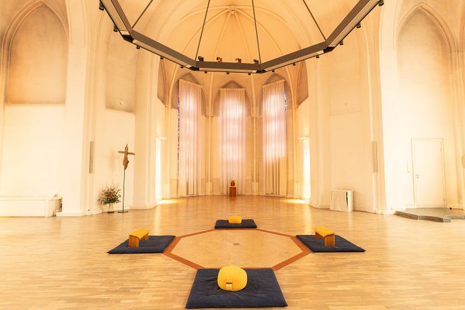 Innenansicht eines hellen Kirchenraums mit Meditationskissen und Bänken auf dem Boden.