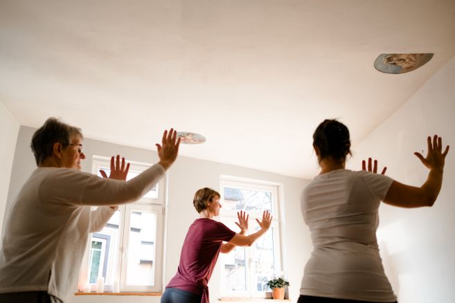 Drei Personen praktizieren Qigong in einem hellen Raum mit Fenstern.