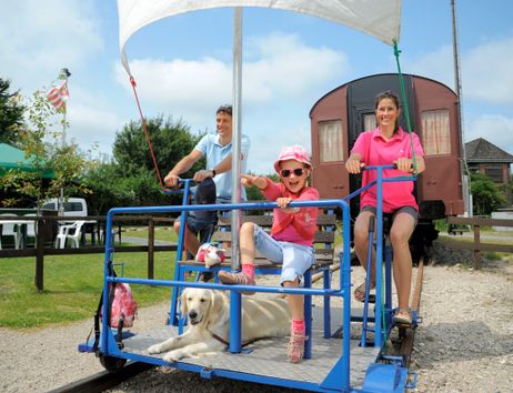 Eine Familie mit einem Hund fährt auf einer Draisine auf Schienen. Im Hintergrund sind ein alter Zugwaggon und ein Garten mit Tischen zu sehen.