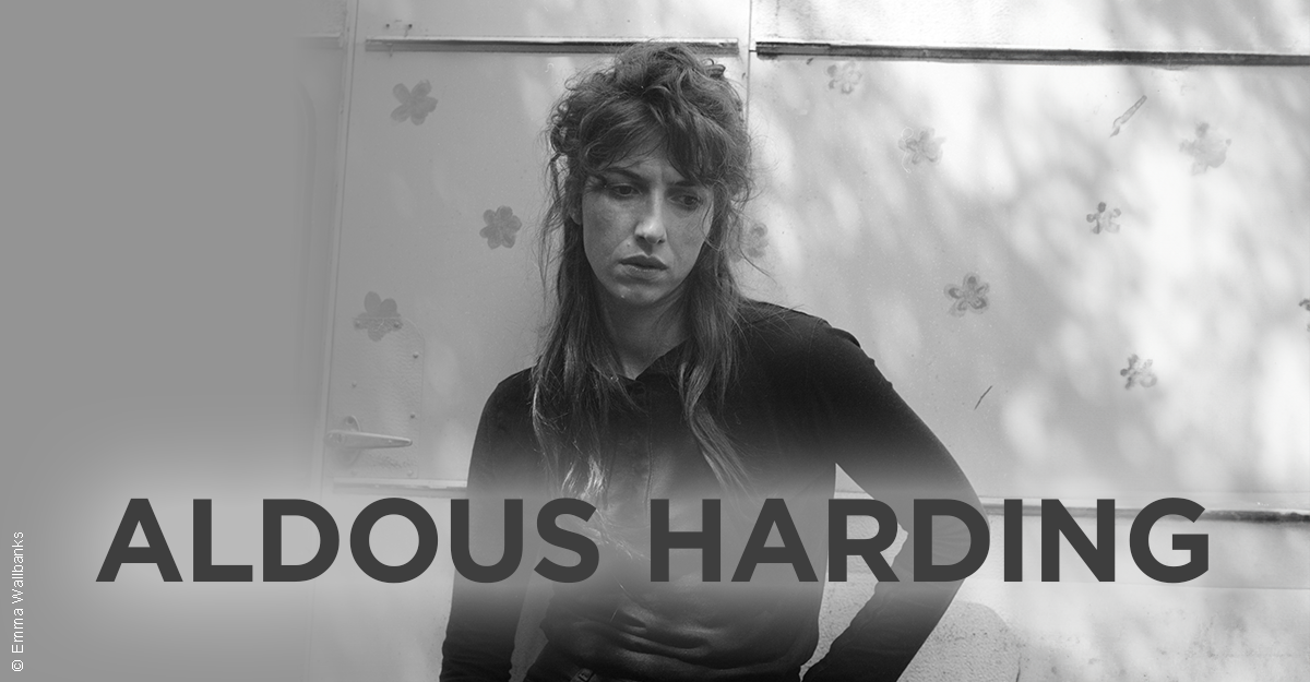 Schwarz-weiß Foto einer Person vor einer Wand mit Blumenmuster. Der Name Aldous Harding ist groß im Vordergrund zu sehen.