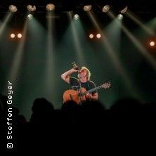 Ein Musiker auf der Bühne mit Gitarre und Mikrofon, umgeben von Scheinwerfern.