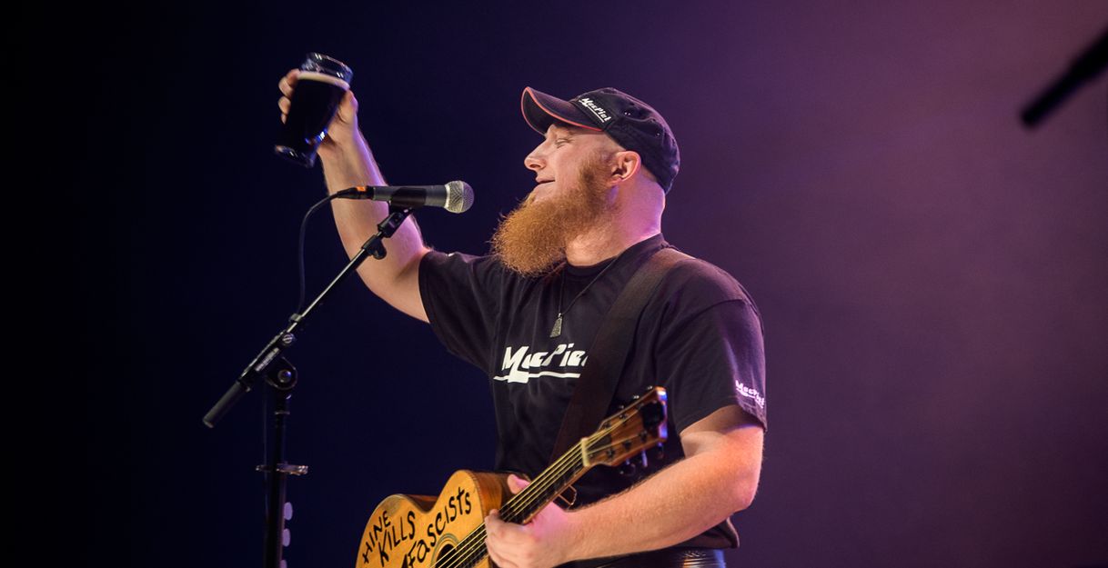 Ein Musiker mit rotem Bart hebt ein Glas auf der Bühne. Er trägt eine schwarze Kappe und ein T-Shirt mit der Aufschrift 'MacPiet'.