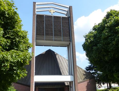 Moderne Kirche mit markantem Turm und Kreuz, umgeben von Bäumen.