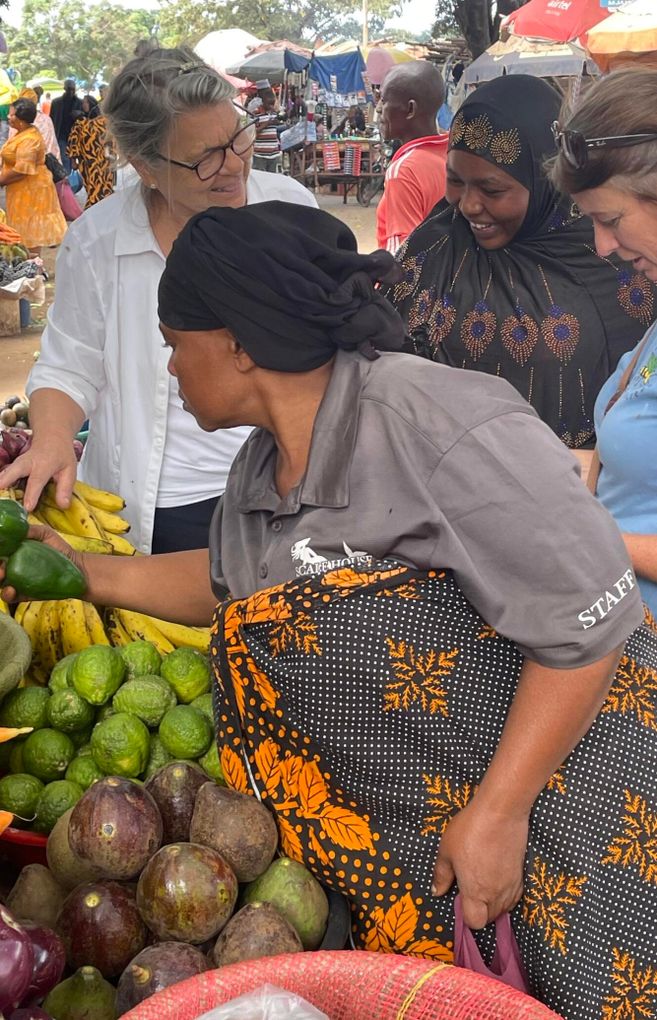 In the picture, people can be seen at a market selling and buying fresh fruits and vegetables. The atmosphere is lively and colorful, and the vendors are wearing traditional clothing.