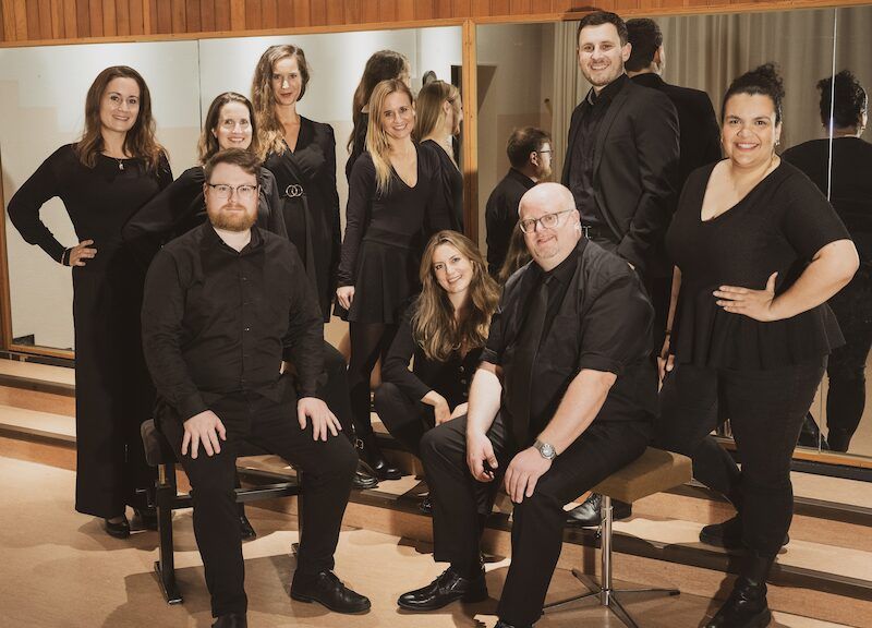 Eine Gruppe von neun Personen in schwarzer Kleidung posiert in einem Raum mit Spiegeln.