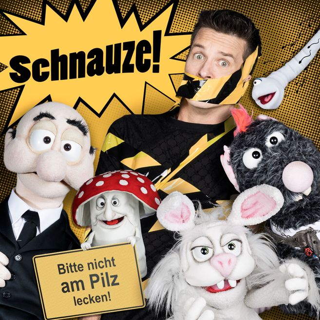 Ein Mann mit Klebeband über dem Mund, umgeben von Puppen, darunter ein Pilz mit einem Schild "Bitte nicht am Pilz lecken!".