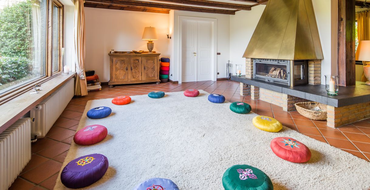 Ein gemütlicher Meditationsraum mit bunten Kissen im Kreis auf einem Teppich, einem Kamin und Holzmöbeln.