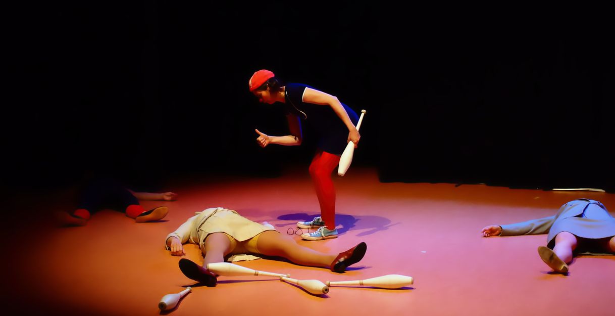 Eine Person in roter Kleidung steht auf einer Bühne mit Jonglierkeulen, umgeben von drei liegenden Personen.