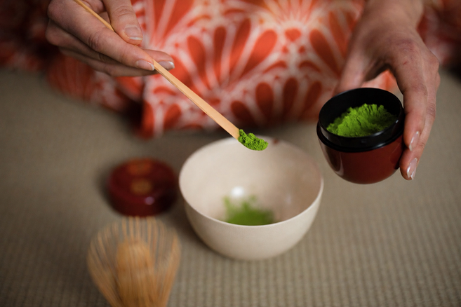 Matcha wird mit einem Bambuslöffel in die Schale gefüllt. Die Frau trägt einen roten Kimono mit weißem Muster drauf.
