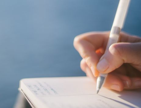 Nahaufnahme einer Hand, die mit einem Stift in ein Notizbuch schreibt, vor einem verschwommenen blauen Hintergrund.