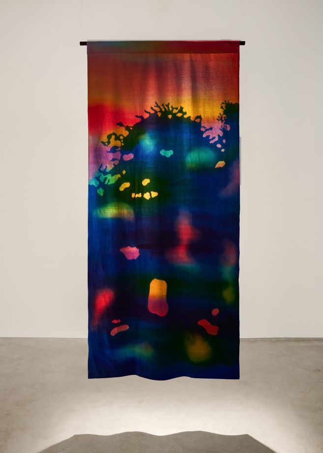 Ein farbenfrohes, vertikal hängendes Textilkunstwerk mit abstrakten Mustern in Rot, Blau, Grün und Gelb.