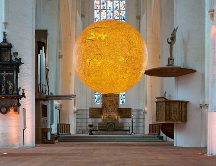 Innenansicht einer Kirche mit einer großen, leuchtenden Kugel, die die Sonne darstellt, über dem Altar.