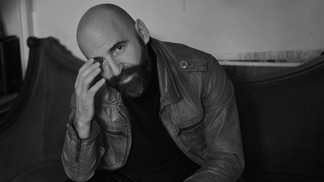 Schwarz-weiß Porträt eines Mannes mit Glatze und Bart, der eine Lederjacke trägt und nachdenklich in die Kamera schaut.