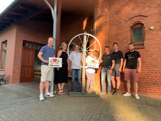 Eine Gruppe von sechs Personen steht vor einem Backsteingebäude neben einem großen Rad mit einem Schild, auf dem 'Herzhorn' steht.