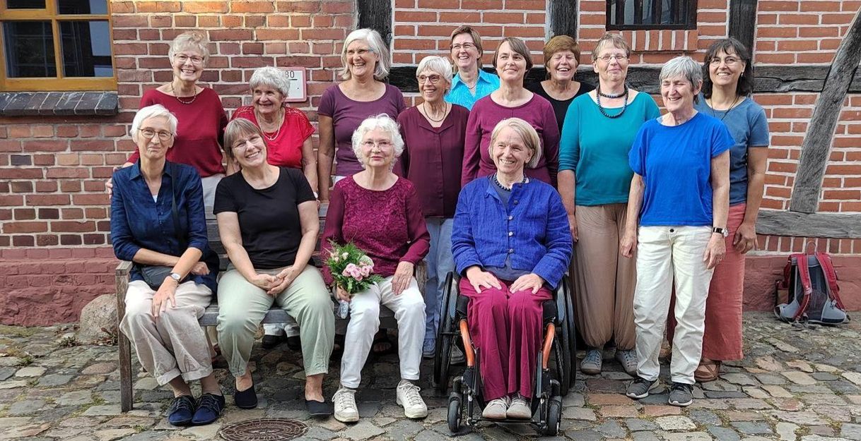 13 Frauen des Chores "FrauenTöne" und Chorleiterin stehen in 2 Reihen vor einem Fahwerkhaus. Sie tragen helle Hosen und Oberteile in Blau- und Rottönen.