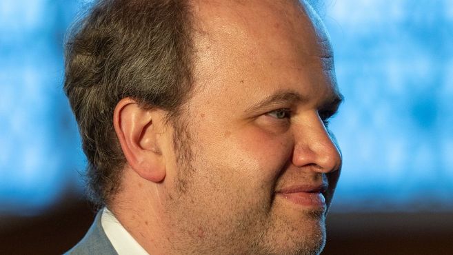 Nahaufnahme eines Mannes mit Bart, der zur Seite schaut, vor einem unscharfen blauen Hintergrund.