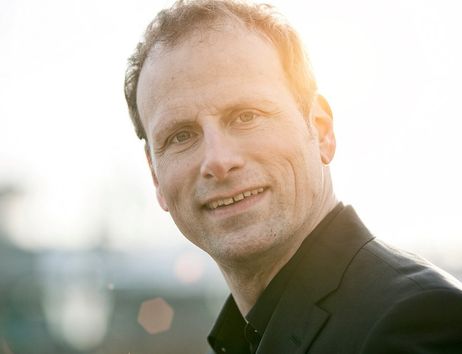 Porträt eines lächelnden Mannes im Anzug vor unscharfem Hintergrund mit Sonnenlicht.