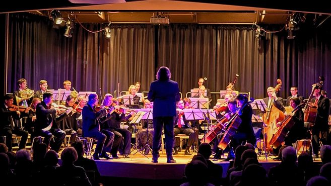 Orchester auf einer Bühne mit Dirigent, Streichern und Bläsern vor Publikum bei einem Konzert.