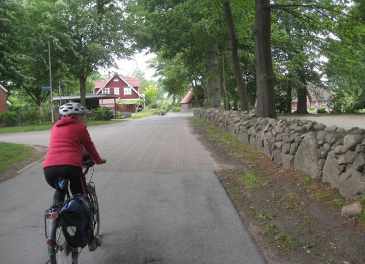 Eine Person fährt mit dem Fahrrad auf einer ländlichen Straße entlang einer Steinmauer, mit Bäumen und einem roten Haus im Hintergrund.