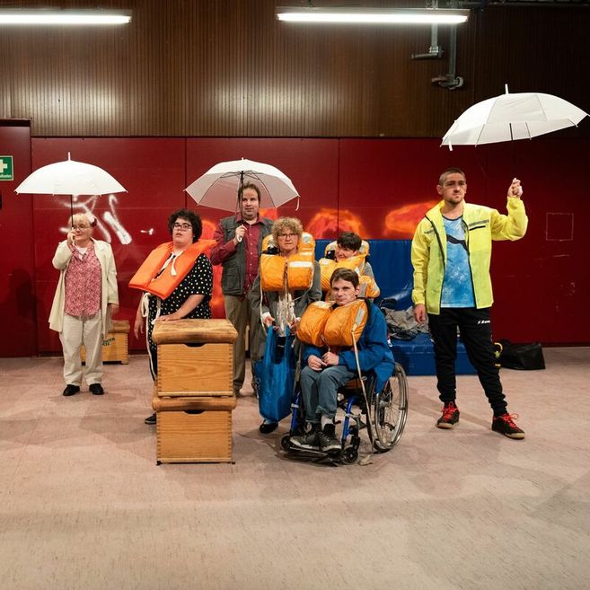 Eine Gruppe von Menschen mit Rettungswesten und Regenschirmen in einem Raum mit roter Wand.
