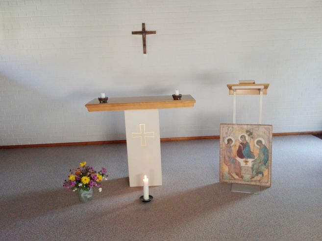 Ein schlichter Altar mit Kreuz, Kerzen, Blumen und einem religiösen Gemälde in einem ruhigen Raum.