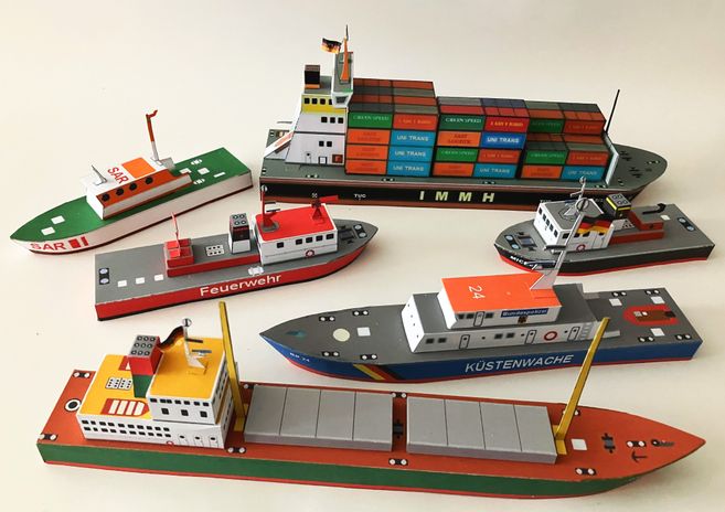 Papiermodelle von verschiedenen Schiffen, darunter ein Containerschiff, ein Küstenwachboot und ein Feuerwehrboot, auf einem Tisch arrangiert.