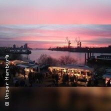 Sonnenaufgang über dem Hamburger Fischmarkt mit Ständen und Hafenkränen im Hintergrund.