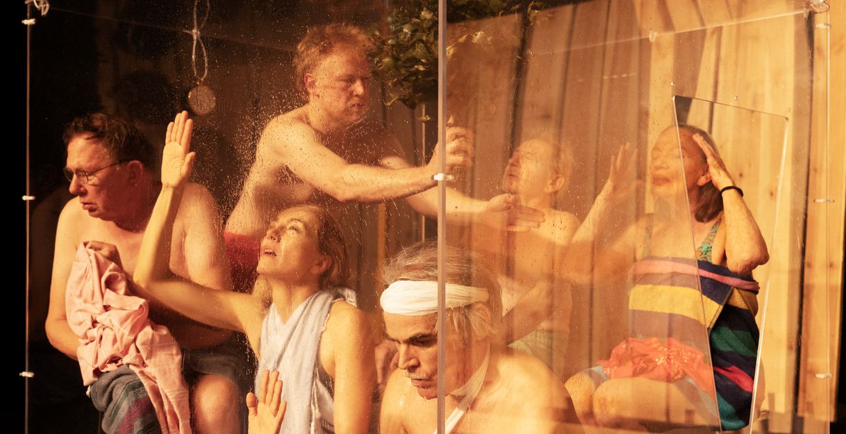 Six performers are in a sauna setting behind a glass wall. They are all in different positions and scantily clad.