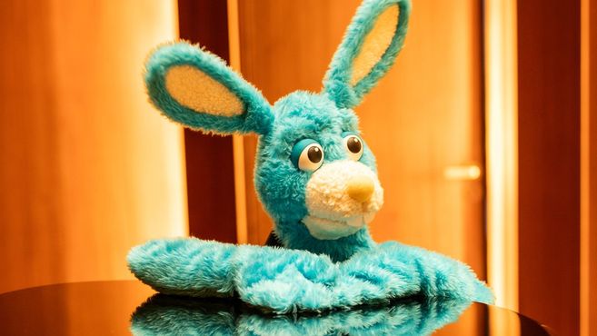 Ein blauer Plüschhase mit großen Augen und langen Ohren sitzt auf einem Tisch vor einem unscharfen Hintergrund.