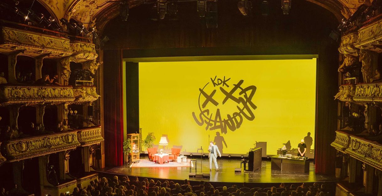 Ein Stand-up-Comedian auf einer Bühne in einem prunkvollen Theater mit gelbem Hintergrund und der Aufschrift 'KDK Stand Up'.