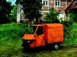 Ein orangefarbenes dreirädriges Fahrzeug steht auf einer grünen Wiese vor einem alten Backsteingebäude.