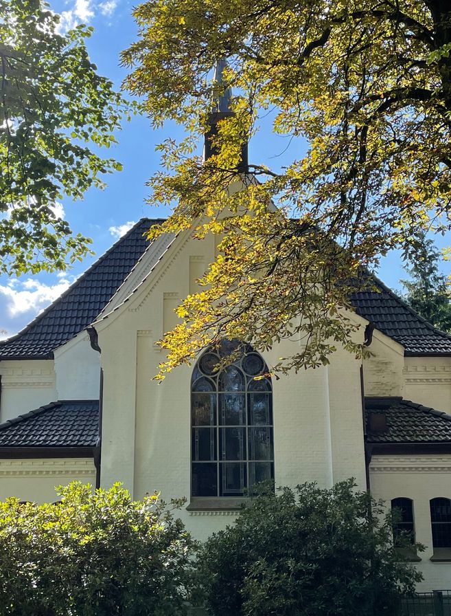 Eine weiße Kirche mit einem großen Fenster und einem schwarzen Dach, umgeben von Bäumen und Sonnenlicht.