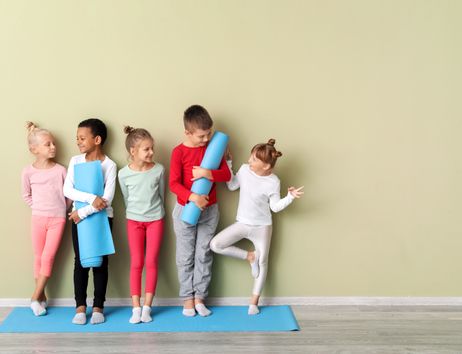Fünf Kinder stehen auf einer blauen Yogamatte vor einer hellgrünen Wand und halten Yogamatten oder machen eine Balance-Übung.