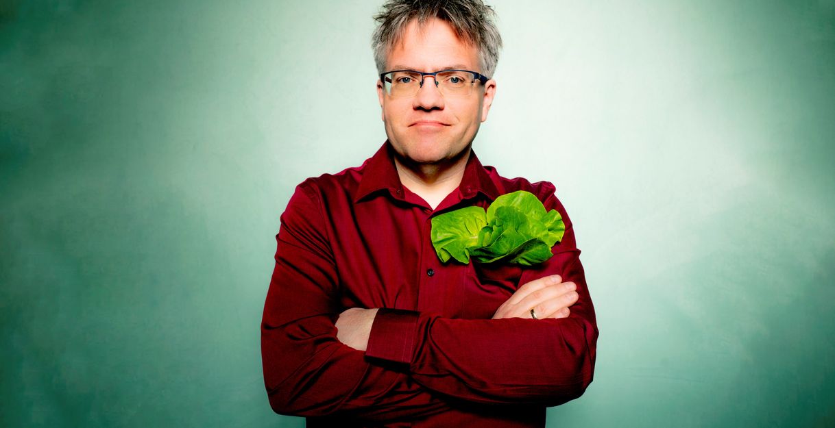 Bild von Matthias Reuter im weinroten Hemd und mit Salatblatt in der Hemdtasche, grüner Hintergrund. 