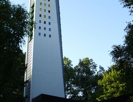 Ein moderner Kirchturm mit bunten Fenstern, umgeben von Bäumen.