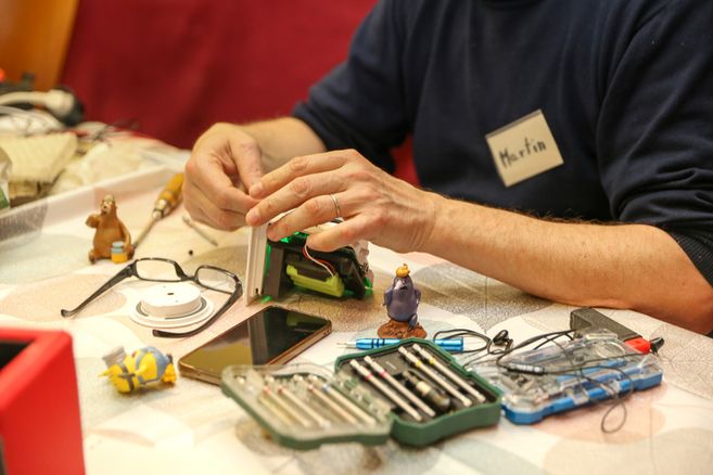 Person repariert ein elektronisches Gerät an einem Tisch mit Werkzeugen und kleinen Figuren.