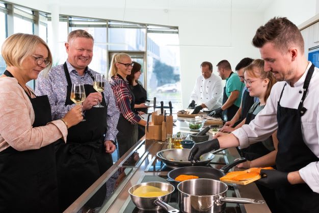 Menschen in einer modernen Küche bei einem Kochkurs, einige kochen, andere trinken Wein.
