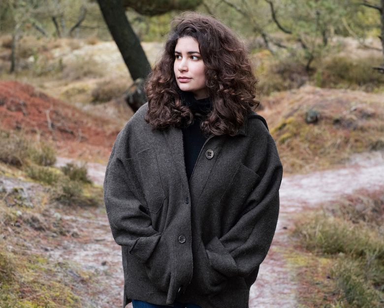 Eine Frau mit lockigem Haar steht in einer herbstlichen Landschaft mit Bäumen und Sträuchern.