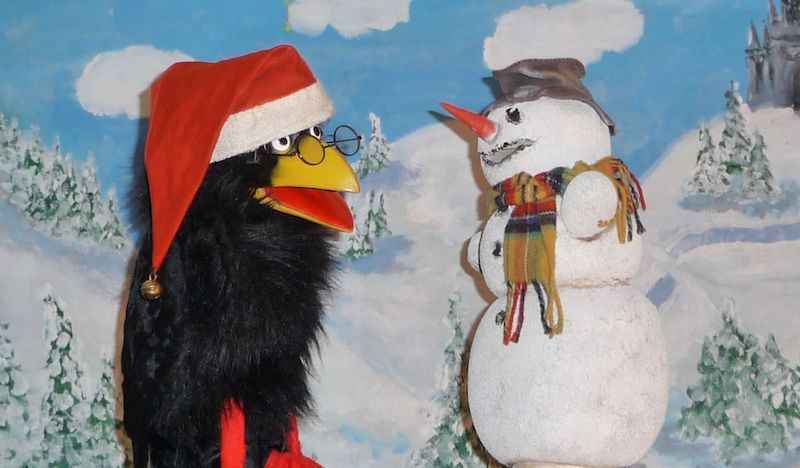 Ein schwarzer Vogel mit roter Mütze und Brille steht vor einem Schneemann mit Hut und Schal vor einer Winterlandschaft.