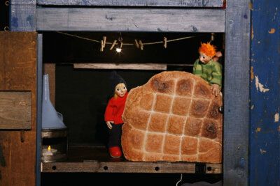 Zwei Puppen mit einem großen Brot in einer Holzkulisse.