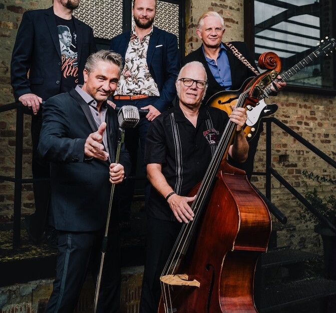 Eine fünfköpfige Band posiert mit Instrumenten vor einem Backsteingebäude.