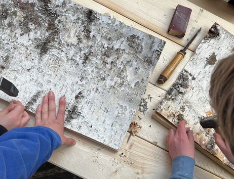 Kinderhände hämmern Nägel in Stücke aus Birkenrinde, daneben liegt Werkzeug (Stecheisen und Schleifpapier)