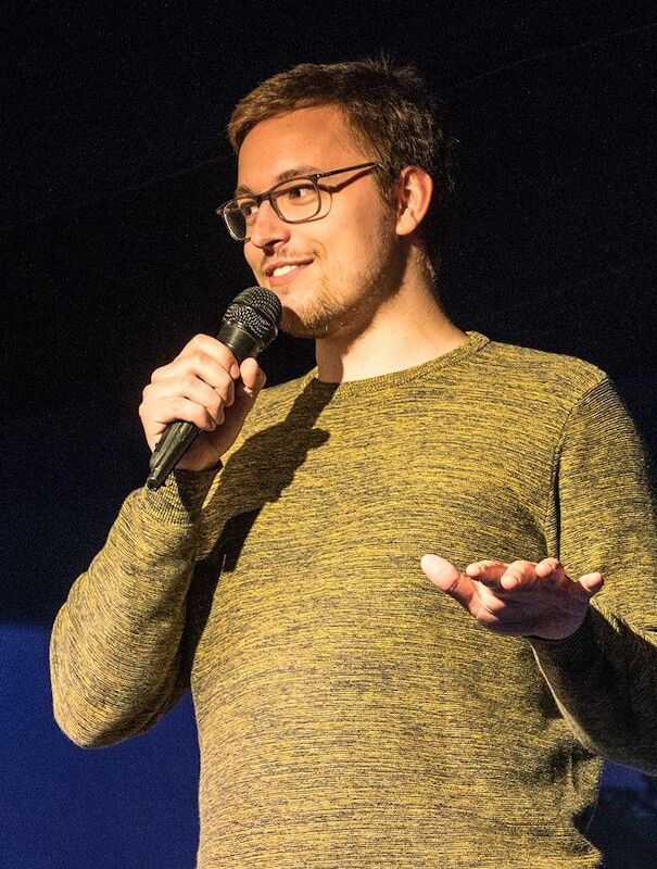 Ein Mann mit Brille und Bart hält ein Mikrofon und spricht auf einer Bühne.