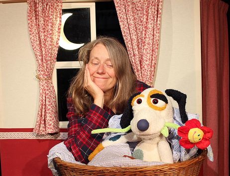 Eine lächelnde Person im Schlafanzug lehnt sich an einen Wäschekorb mit einem kuscheligen Stoffhund, der eine Blume im Maul hält. Im Hintergrund sind ein Fenster mit Mond und gemusterte Vorhänge zu sehen – eine gemütliche Schlafzimmerszene.