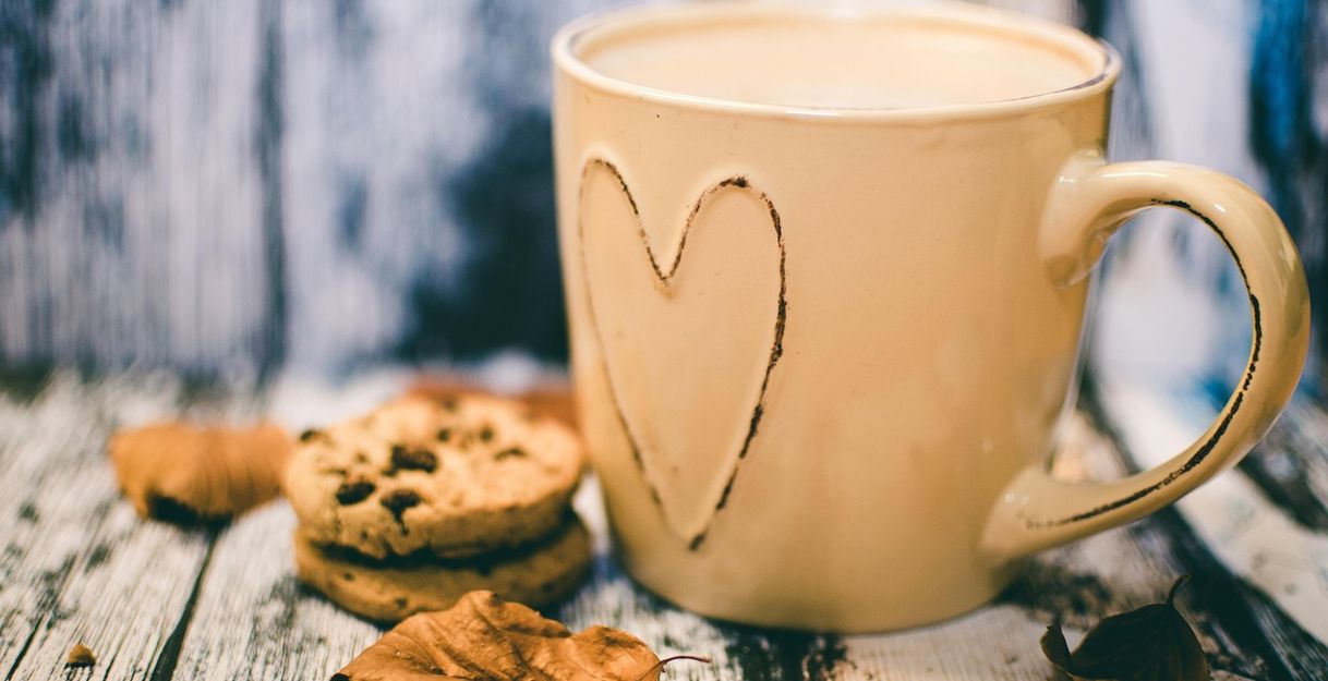 Eine beige Tasse mit Herzmotiv, Kekse und Herbstblätter auf einem Holztisch.