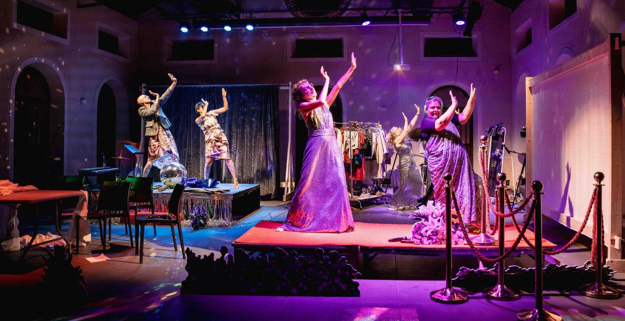 The performers dance around the stage to Bollywood music, making big gestures. There are colorful lights and the glow of a disco ball.